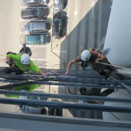 Dwóch alpinistów przemysłowych w kaskach i uprzężach czyści szklaną fasadę budynku biurowego, widok z góry na parking z samochodami.
