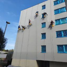 Grupa alpinistów przemysłowych, zabezpieczonych linami, maluje elewację budynku mieszkalnego; okna zabezpieczone folią, widoczne wiaderka z farbą i wałki.