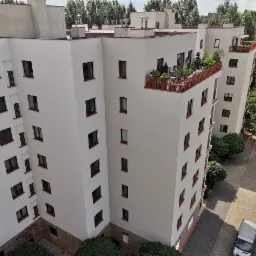 Widok z góry na biały blok mieszkalny z balkonami, zieleń na balkonach, mały samochód dostawczy na parkingu, drzewa w tle.