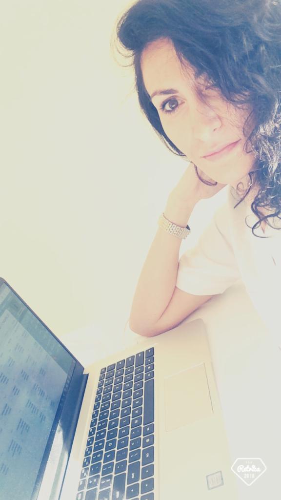 Kobieta z ciemnymi kręconymi włosami opiera się na laptopie, na ekranie widoczne dane, na nadgarstku srebrna bransoletka, uśmiech w oczach, selfie z góry w jasnym otoczeniu.