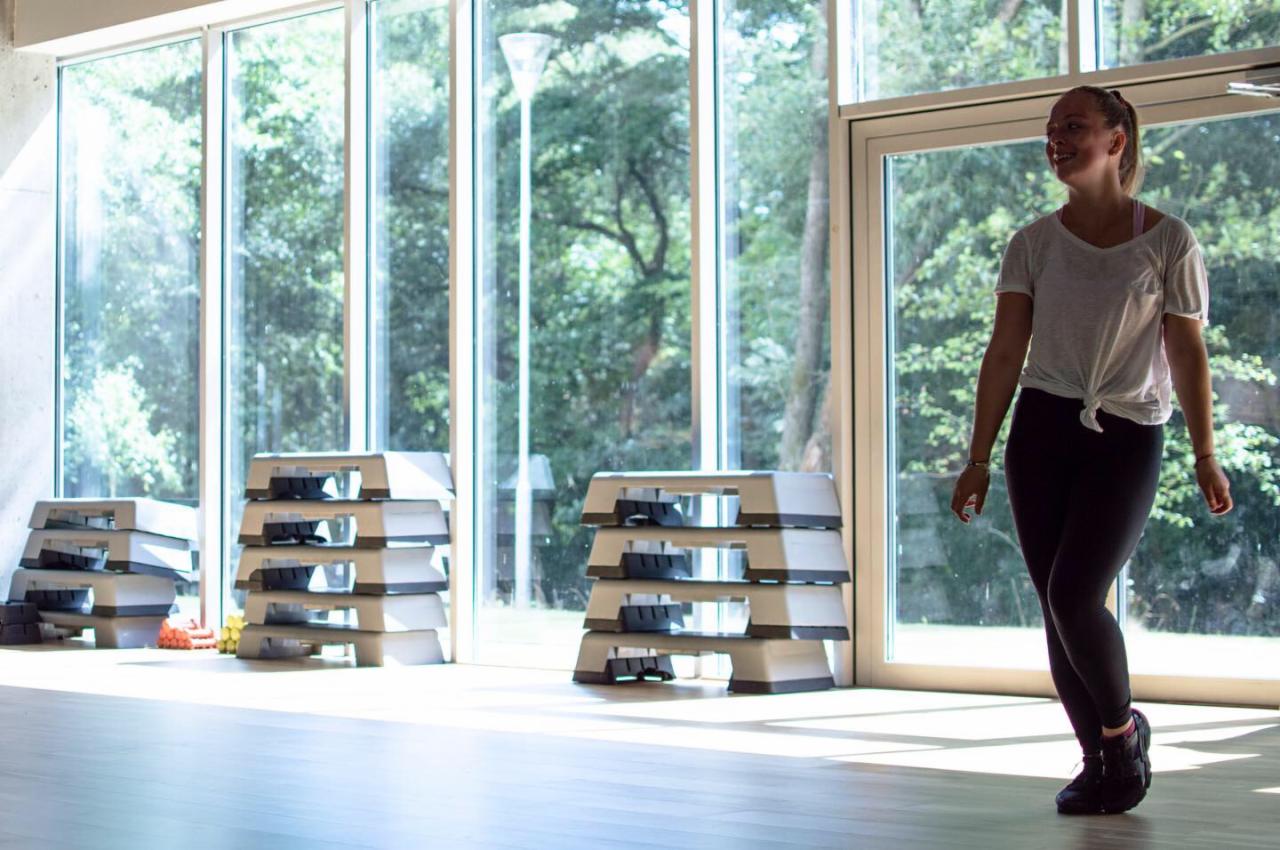 Kobieta w sportowym stroju w jasnym pomieszczeniu z dużymi oknami, obok ułożone w stosy stepy do ćwiczeń.