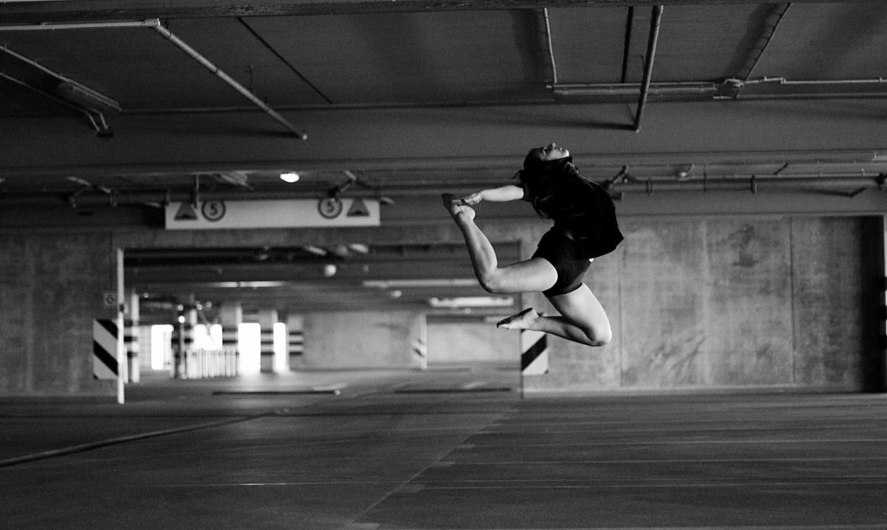 Kobieta w czarnym stroju w dynamicznym skoku, wykonująca element taneczny na tle surowego, betonowego parkingu; czarno-biała fotografia.