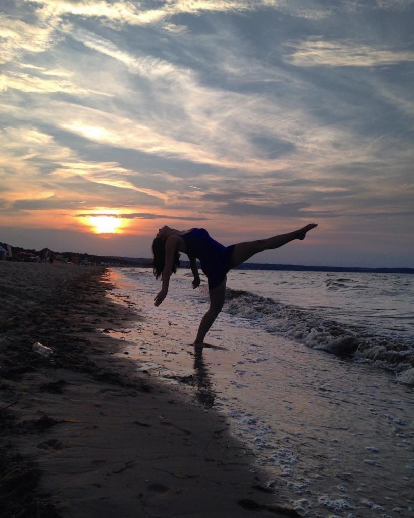 Sylwetka tancerki w granatowym stroju, wykonującej figurę na plaży o zachodzie słońca, z odbiciem w mokrym piasku.
