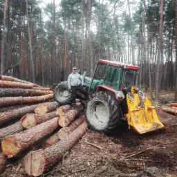 Mężczyzna oparty o traktor z żółtym pługiem na tle sterty ściętych pni drzew w lesie. Widoczne tablice rejestracyjne traktora i wysokie drzewa w tle.