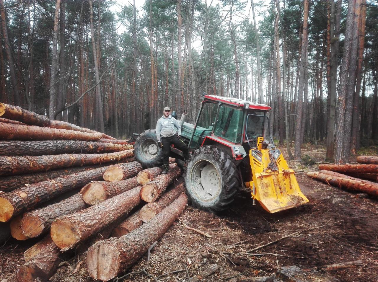 Mężczyzna oparty o traktor z żółtym pługiem na tle sterty ściętych pni drzew w lesie. Widoczne tablice rejestracyjne traktora i wysokie drzewa w tle.