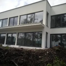 Nowoczesny dom z dużymi oknami w ciemnych ramach, widoczne prace ziemne wokół budynku.