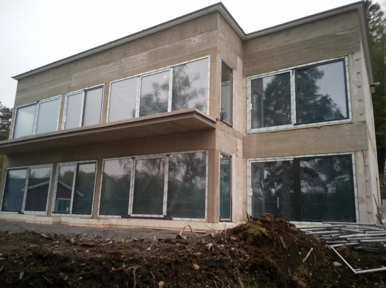 Surowy, dwukondygnacyjny budynek w trakcie budowy, z widocznymi otworami okiennymi z częściowo zamontowanymi oknami, betonowa konstrukcja w stanie niedokończonym, widoczne rusztowania i ziemia.