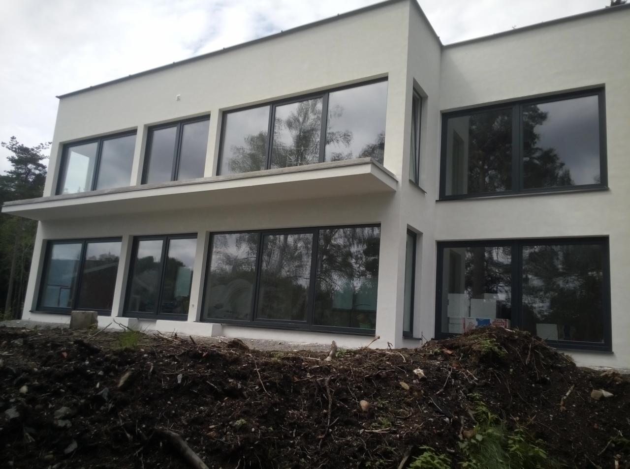 Nowoczesny dom z dużymi oknami w ciemnych ramach, widoczne prace ziemne wokół budynku.
