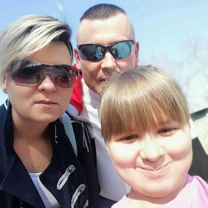 Selfie rodziny: kobieta w kurtce z ozdobnymi guzikami, mężczyzna w okularach i uśmiechnięta dziewczynka na tle błękitnego nieba. Jasne oświetlenie.