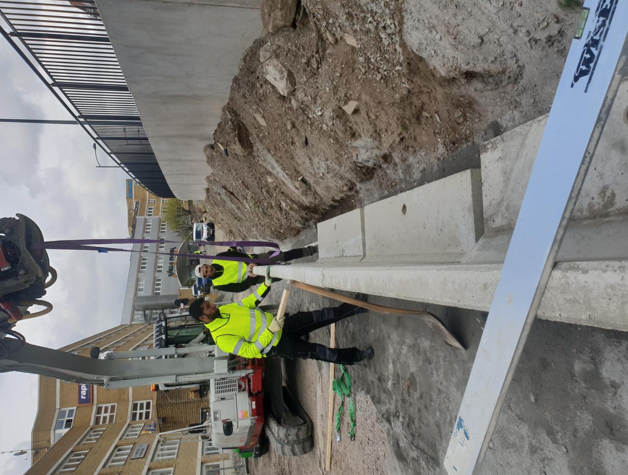 Dwóch pracowników w żółtych kamizelkach montuje betonowe elementy fundamentu przy użyciu koparki i łopaty, widoczny wykop i budynek w tle.