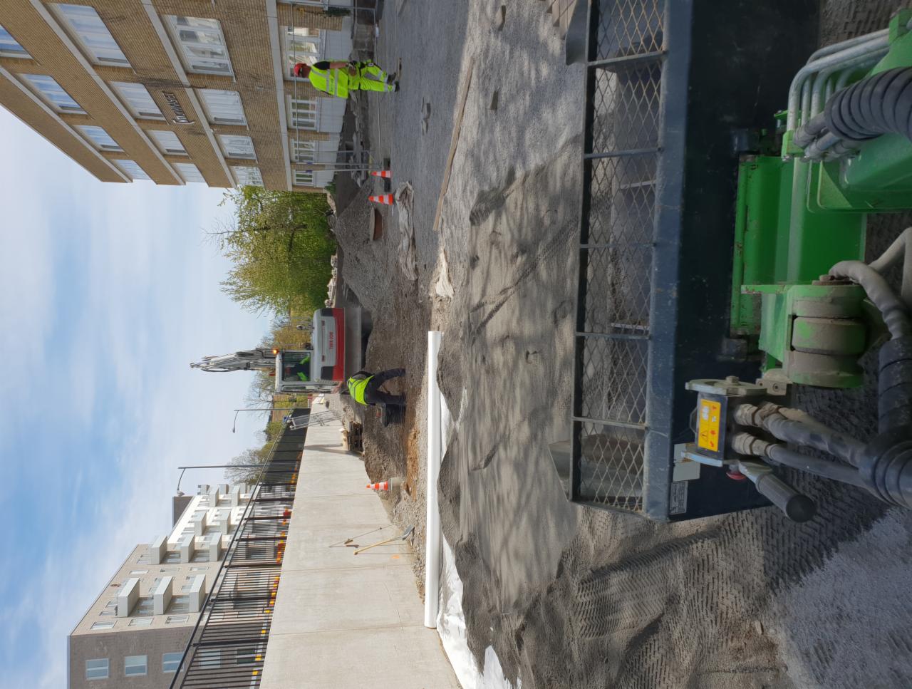 Prace ziemne z użyciem koparki na placu budowy, widoczni pracownicy w odblaskowych kamizelkach i rury drenażowe.