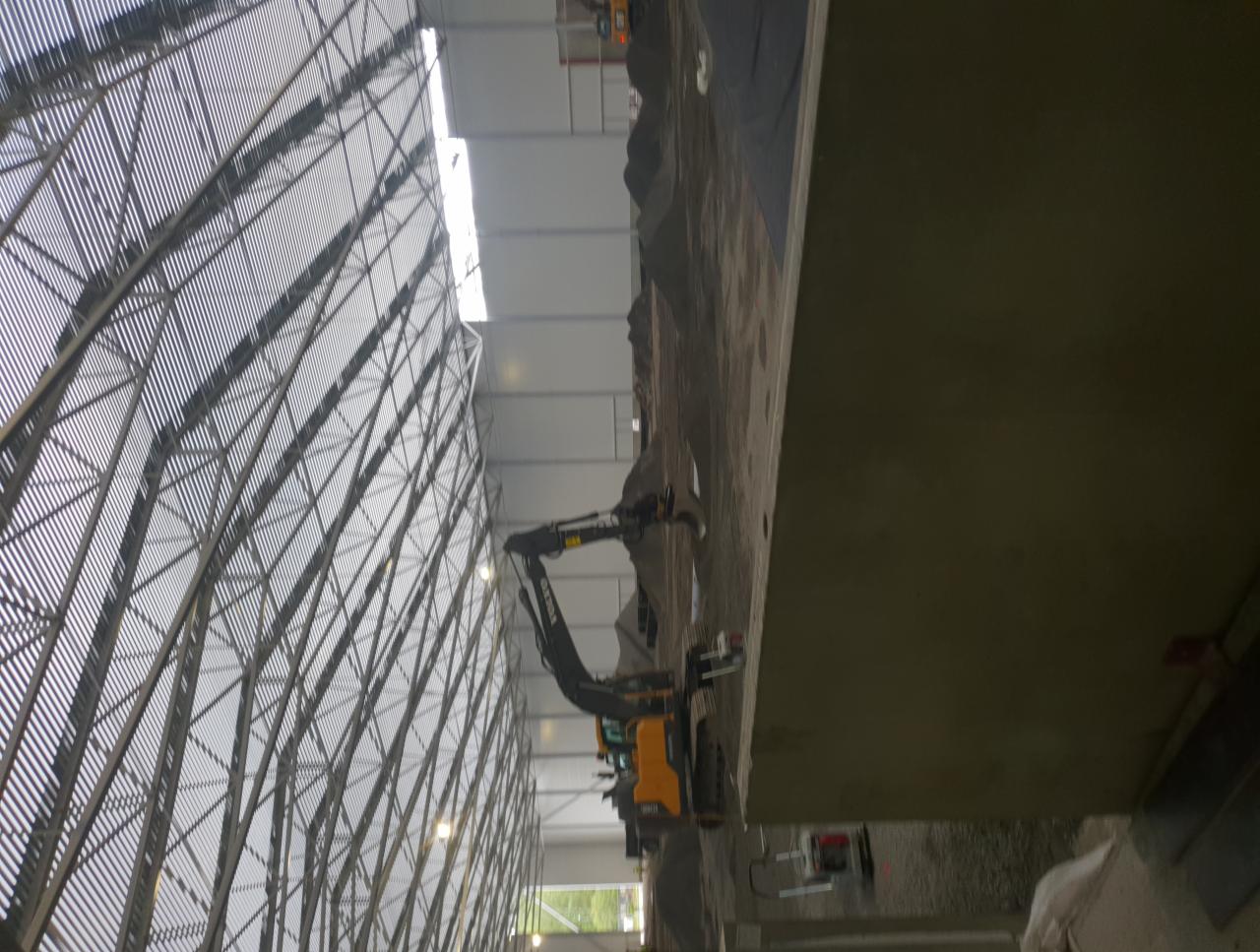 Żółta koparka pracuje wewnątrz hali o konstrukcji stalowej, usypując ziemię.