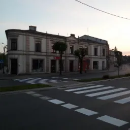 Dwupiętrowy budynek z jasną elewacją i ciemną stolarką okienną widziany z perspektywy ulicy z przejściem dla pieszych wczesnym rankiem.
