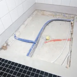 Przygotowane podłoże pod prysznic z widocznymi rurami instalacji wodnej i kanalizacyjnej, czarna mozaika na obrzeżach, białe płytki na ścianie.