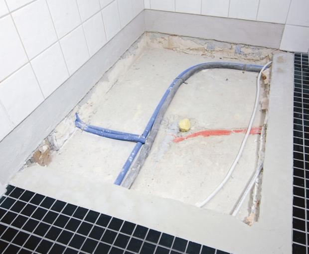 Przygotowane podłoże pod prysznic z widocznymi rurami instalacji wodnej i kanalizacyjnej, czarna mozaika na obrzeżach, białe płytki na ścianie.