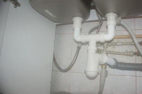 Widok od dołu na podwójny zlew kuchenny z widocznymi białymi rurami odpływowymi z tworzywa sztucznego, syfonem i przyłączami do instalacji wodnej na tle białych płytek.