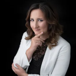 Portret kobiety w białej marynarce i czarnej koronkowej bluzce, z palcem wskazującym dotykającym brody, na czarnym tle.