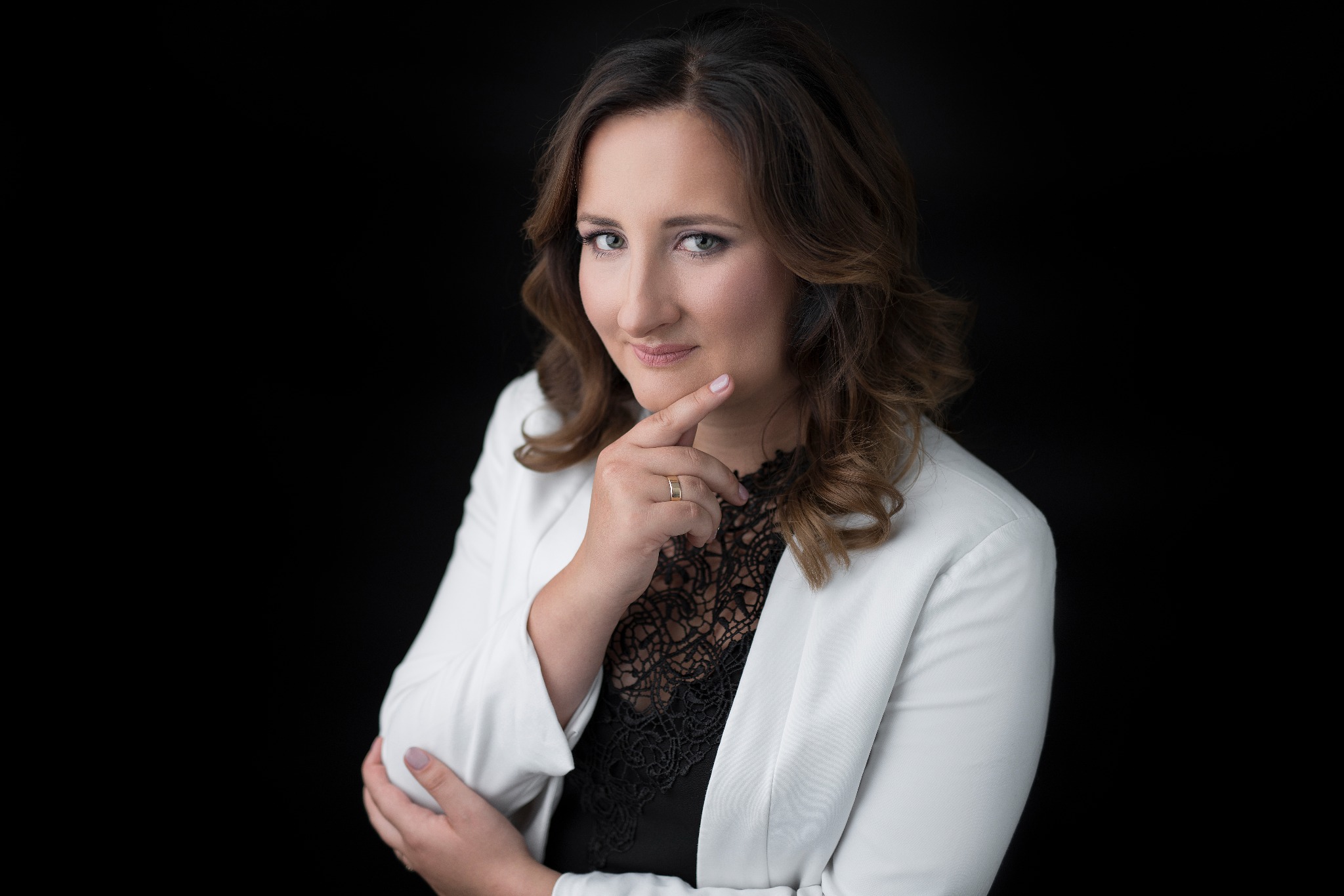Portret kobiety w białej marynarce i czarnej koronkowej bluzce, z palcem wskazującym dotykającym brody, na czarnym tle.