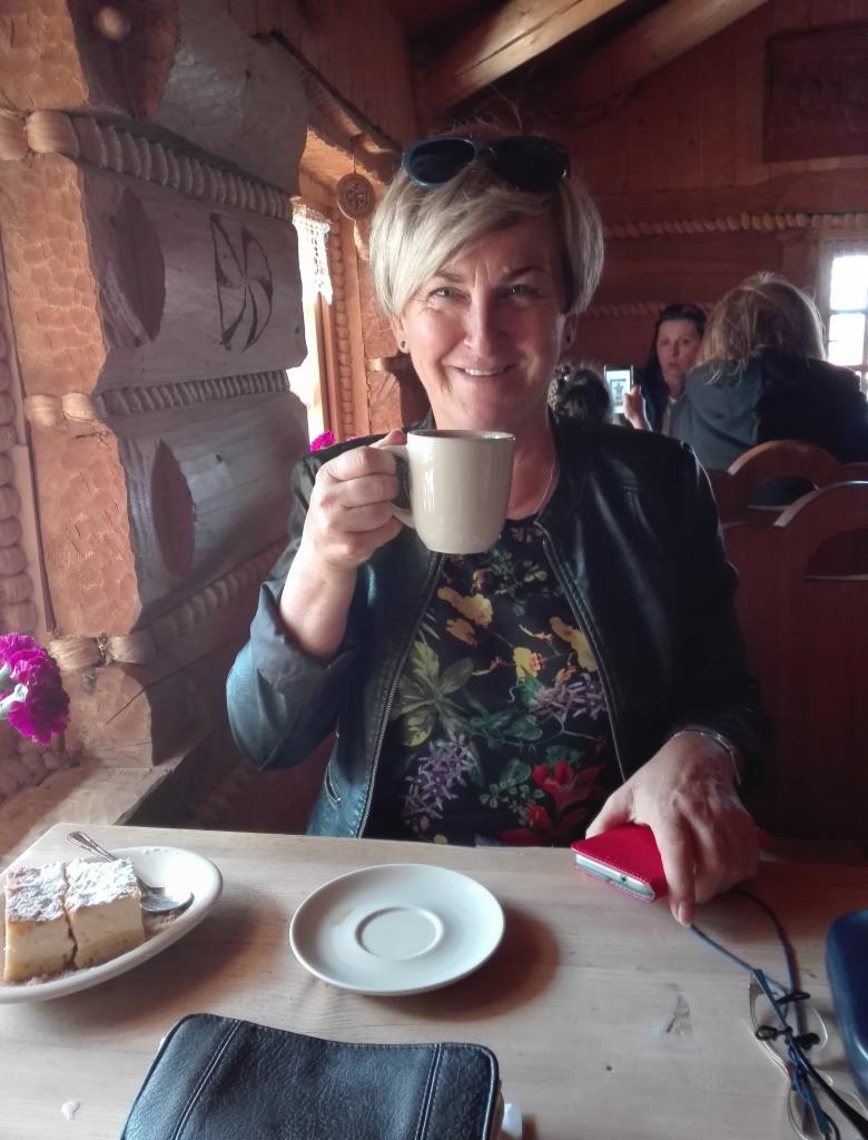 Uśmiechnięta kobieta z krótkimi blond włosami, w skórzanej kurtce i kwiecistej bluzce, trzyma filiżankę z napojem w drewnianym wnętrzu, na stole ciasto z cukrem pudrem, torebka i etui na okulary.