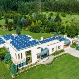 Rozległa posiadłość z białym domem i licznymi panelami słonecznymi na dachu, otoczona bujną zielenią, basenem i stawem.