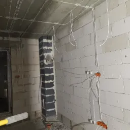 Surowe wnętrze w trakcie prac elektrycznych: widoczne przewody rozprowadzone po ścianach z betonu komórkowego i suficie, pomarańczowe puszki instalacyjne pod tynk oraz fragment kolumny...