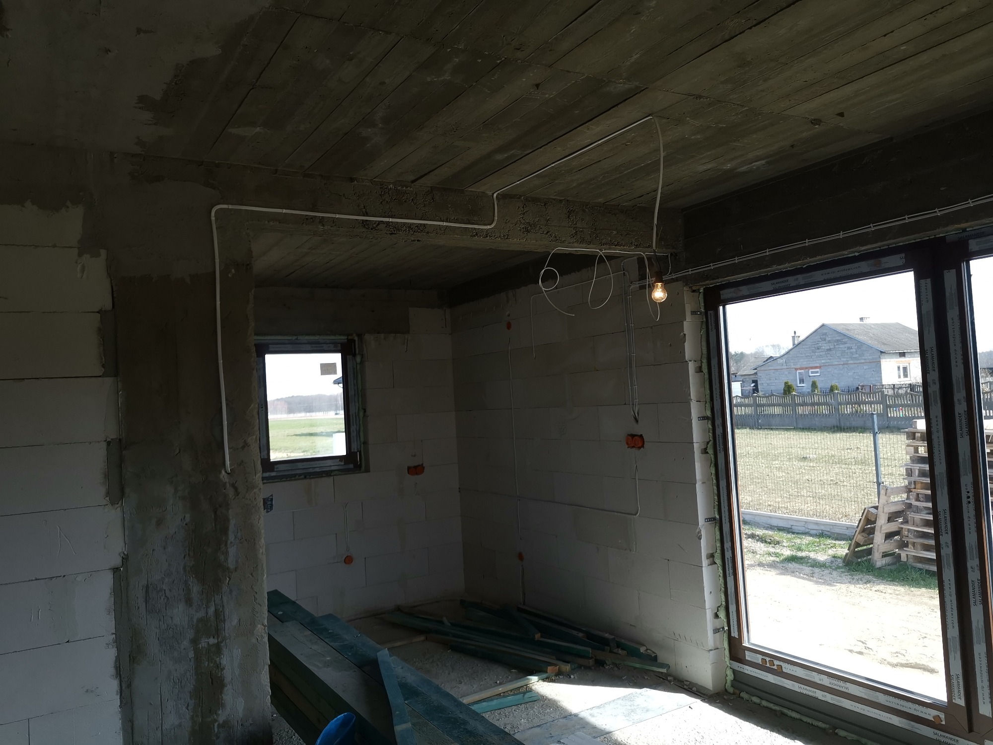 Surowy stan wnętrza domu w trakcie budowy, widoczne poprowadzone przewody elektryczne pod sufitem, gniazdka w ścianach z betonu komórkowego oraz prowizoryczne oświetlenie żarówką.