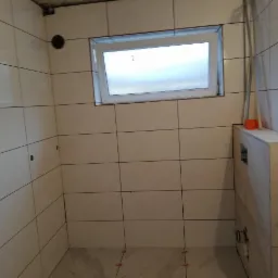 Świeżo wykafelkowana łazienka z małym oknem, widoczne krzyżyki dystansowe między płytkami na podłodze, częściowo zabudowany stelaż podtynkowy WC, białe płytki na ścianach z ciemną fugą.