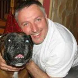 Uśmiechnięty mężczyzna w białej koszulce obejmuje boksera z widoczną bielmem na jednym oku. Tło rozmyte, wnętrze mieszkania.