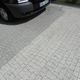 Częściowo wyczyszczona kostka brukowa w jasnoszarym odcieniu, z widocznym fragmentem samochodu w lewym górnym rogu.