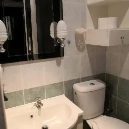 Umywalka z baterią, toaleta i lustro z oświetleniem w odnowionej łazience z szaro-zielonymi płytkami.