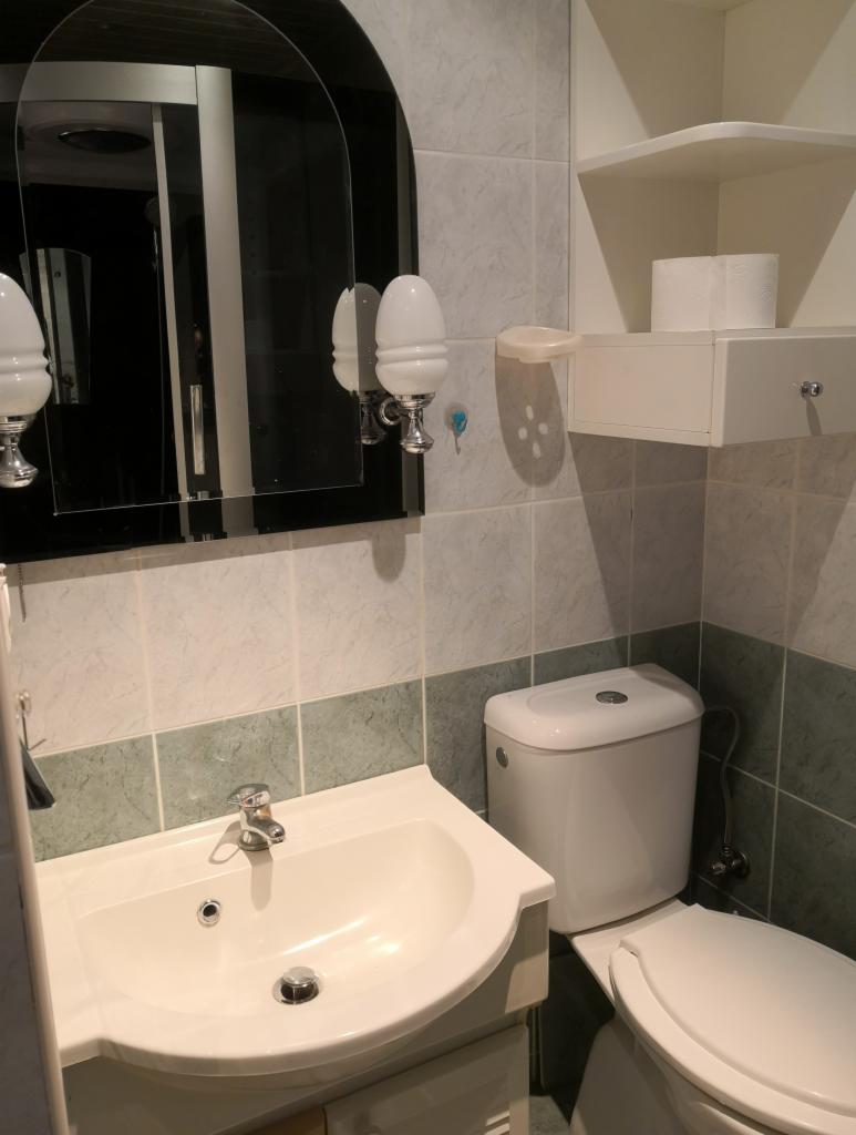 Umywalka z baterią, toaleta i lustro z oświetleniem w odnowionej łazience z szaro-zielonymi płytkami.