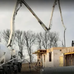 Betonomieszarka CIFA z wysięgnikiem pompuje beton na budowę domu z bloczków, widoczni pracownicy w ubraniach roboczych, drzewa bez liści w tle.