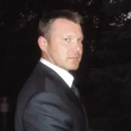 Portret mężczyzny w garniturze, patrzącego przez ramię, na ciemnym tle z zielenią. Ujęcie od ramion w górę, z delikatnym uśmiechem i krótkimi włosami.