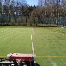 Maszyna czyszcząca sztuczną trawę na boisku sportowym, widoczne linie boiska i siatka ogrodzeniowa w tle.