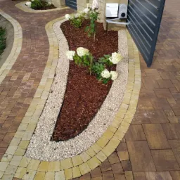 Kaskadowy ogródek z białymi hortensjami, wysypany korą i obłożony białym żwirem, przylegający do ścieżki z kostki brukowej w odcieniach brązu i beżu, z automatyczną bramą w tle.