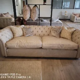 Beżowa sofa Chesterfield z poduszkami w salonie, widoczna z przodu, z pikowanym oparciem i podłokietnikami, na drewnianej podłodze i dywanie.