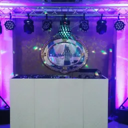 Stanowisko DJ-a z białym panelem frontowym, oświetleniem scenicznym i lustrem w złotej ramie odbijającym salę balową z dekoracjami.