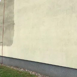 Fragment elewacji budynku z widocznym śladem po czyszczeniu, kontrastującym z zabrudzoną powierzchnią; drabina oparta o ścianę, trawnik u podnóża ściany.