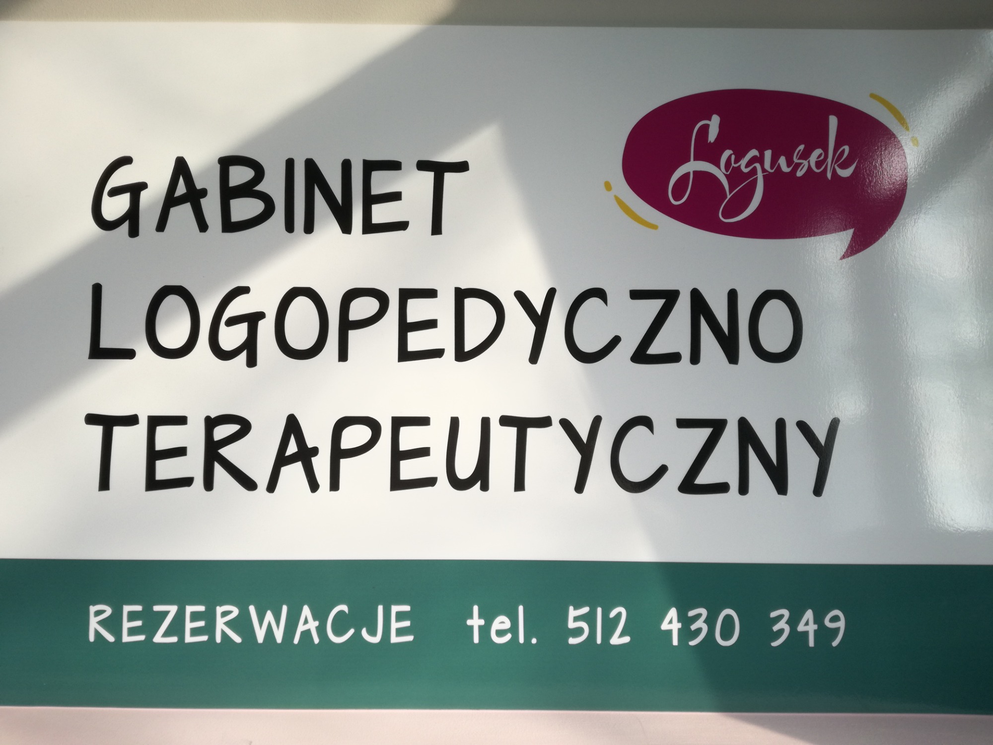 Szyld gabinetu logopedyczno-terapeutycznego z napisem 'Gabinet Logopedyczno Terapeutyczny' w kolorze czarnym, logo w kształcie dymku z napisem 'Logusek' w kolorze różowym oraz numerem telefonu...