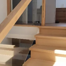 Jasne, drewniane schody z balustradą ze szkła i drewna, w tle widoczne elementy wykończenia wnętrza.