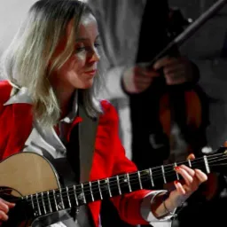 Kobieta w czerwonej marynarce gra na gitarze akustycznej, za nią zamazana postać z skrzypcami, w tle stonowane kolory.