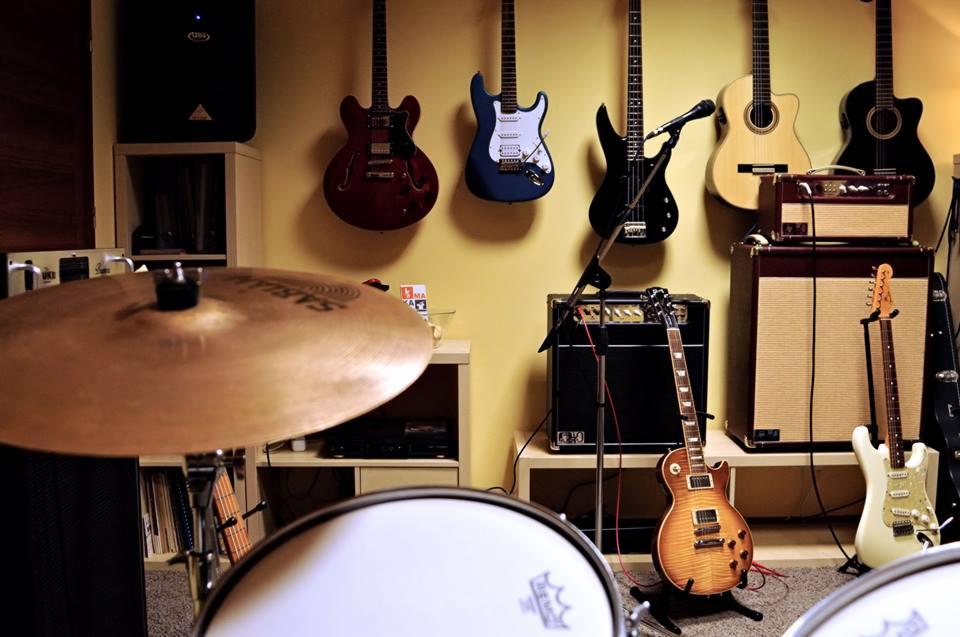 Studio muzyczne z zestawem perkusyjnym na pierwszym planie i kolekcją gitar wiszących na ścianie, w tym gitara basowa i akustyczna, oraz wzmacniacze.