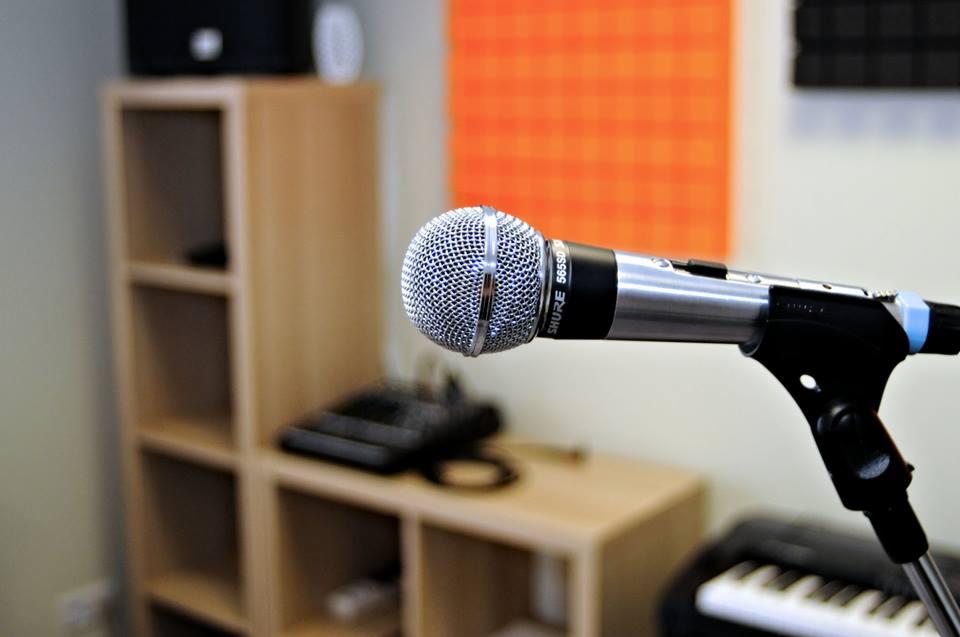 Mikrofon Shure 565SD na statywie w domowym studiu nagraniowym, w tle półka z akcesoriami i panel akustyczny.