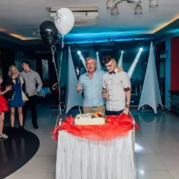 Uroczyste przyjęcie: mężczyźni wznoszą toast nad tortem urodzinowym, w tle goście i dekoracje świetlne.