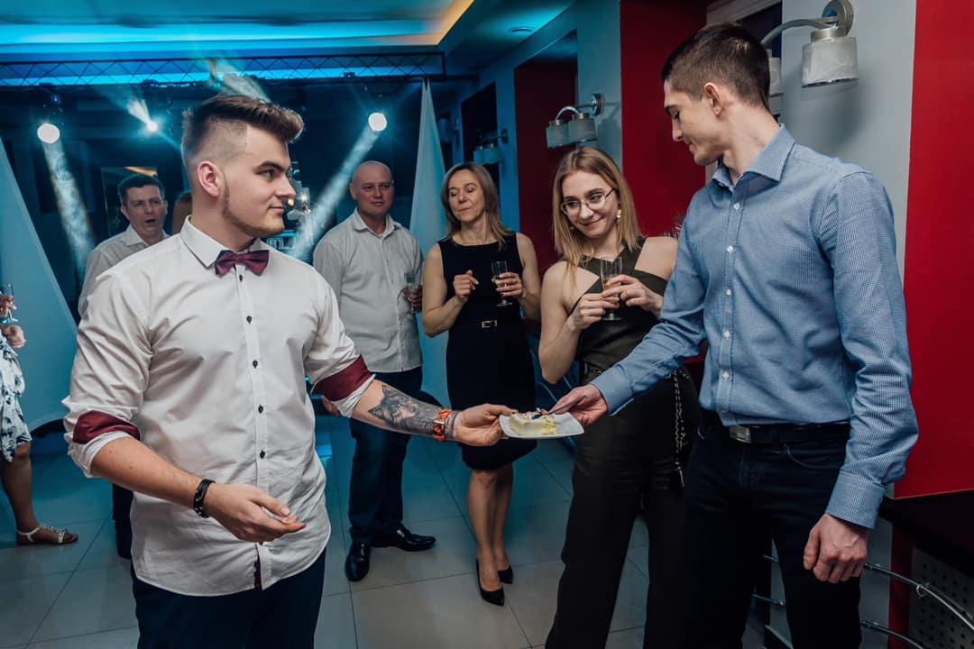 Mężczyzna w białej koszuli z bordową muszką podaje kawałek tortu innemu mężczyźnie na przyjęciu, w tle goście z kieliszkami w dłoniach.