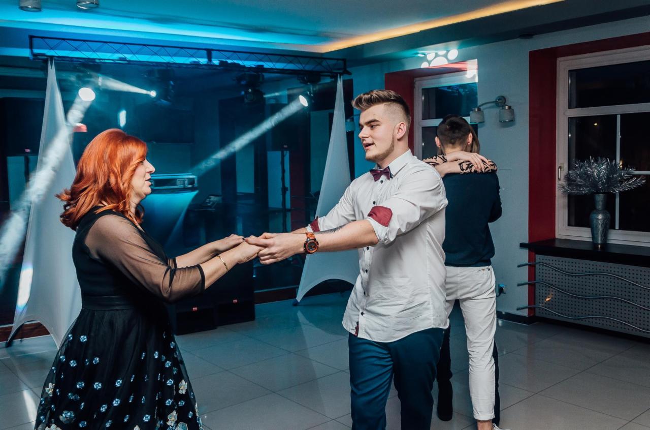 Para trzymająca się za ręce na sali balowej, oświetlenie sceniczne w tle, widoczna para w tle.