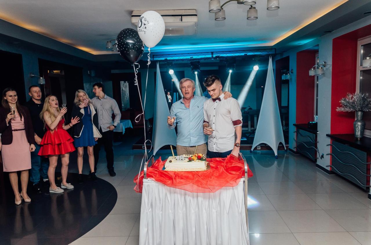 Uroczyste przyjęcie: mężczyźni wznoszą toast nad tortem urodzinowym, w tle goście i dekoracje świetlne.