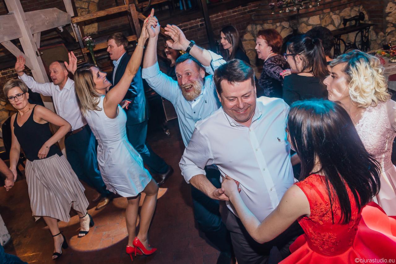 Rozbawieni goście weselni tańczą na parkiecie, kobieta w białej sukience i czerwonych szpilkach unosi ręce, w tle mur z kamienia.