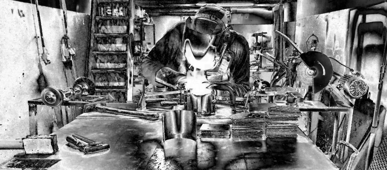 Monochromatyczne ujęcie spawacza w masce ochronnej, wykonującego pracę przy stole warsztatowym z narzędziami i stosem metalowych elementów.