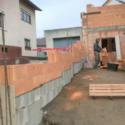 Budowa domu jednorodzinnego: murarz w szarej bluzie układa bloczki ceramiczne na fundamencie z betonu komórkowego, widoczny poziomica laserowa, w tle fragment konstrukcji domu w stanie surowym...